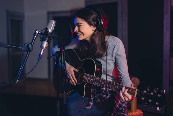 Female musician playing guitar and smiling while recording vocals in studio session