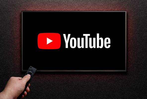 Hand holding remote pointed at TV screen displaying YouTube logo on dark wall background