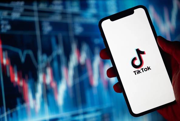 Hand holding smartphone displaying TikTok logo with blurred stock market chart background