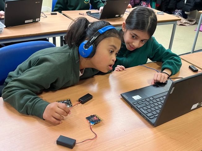 Two pupils look at their computer screen, the girl on the left has headphones on and is listening intently to the tune made by the micro:bit