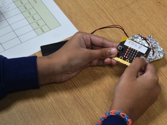 A child holds a micro:bit that has 'Foil 8' written on it, while another micro:bit is wrapped in foil