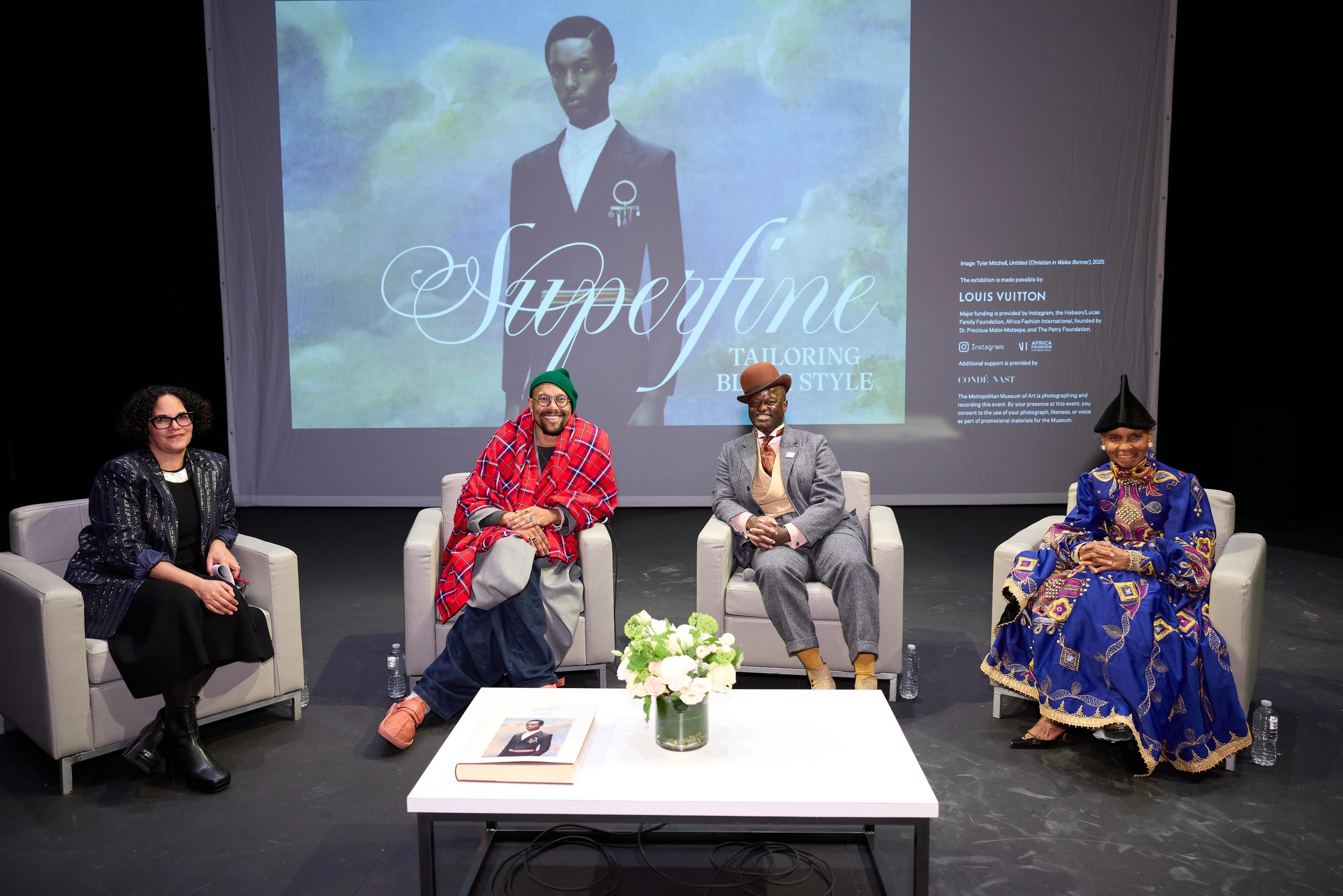 Four individuals are seated on stage in a panel discussion. Behind them, a screen shows "Superfine Tailoring Black Style" and an elegant fashion image. The setting appears formal and thoughtful.