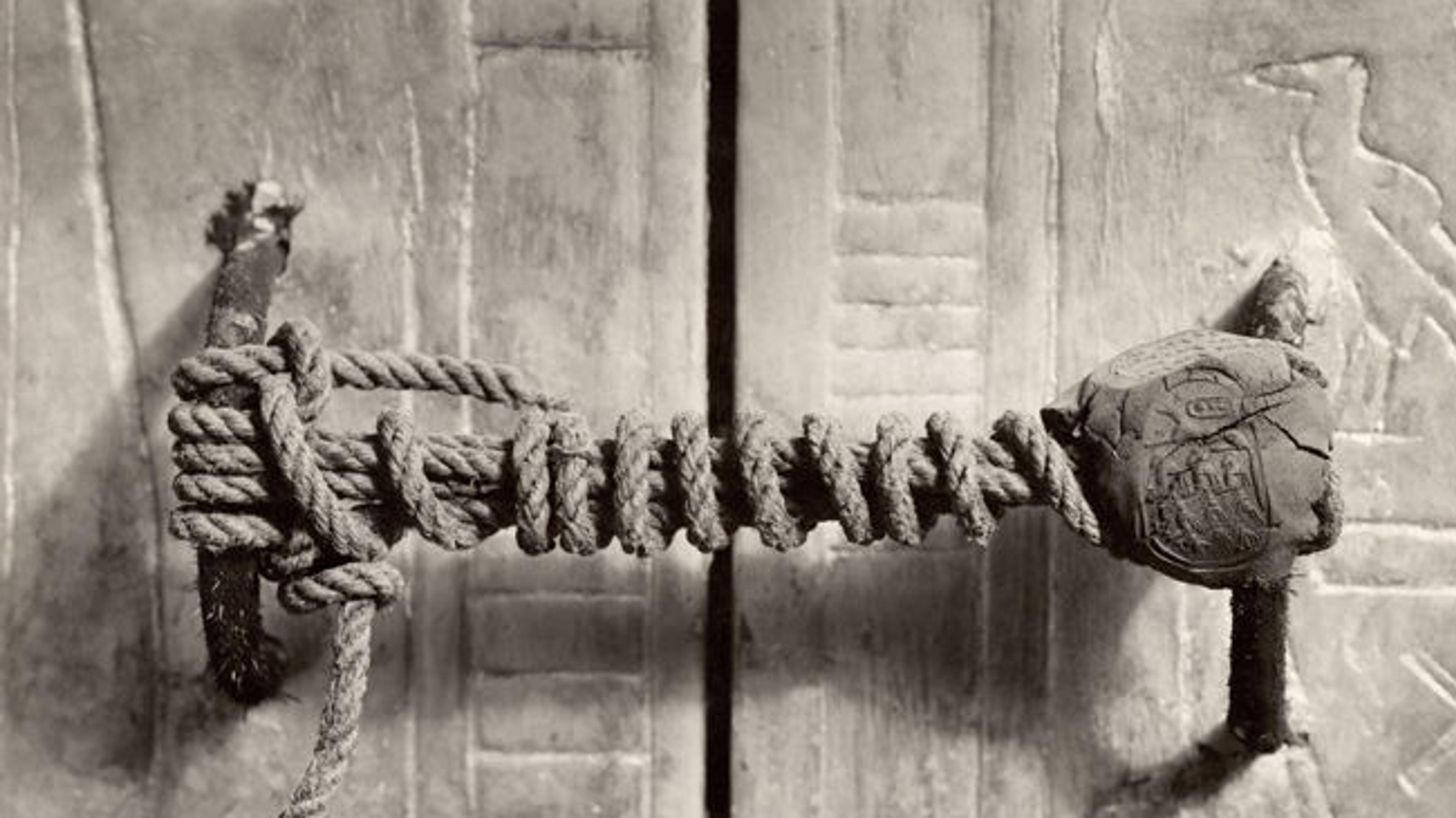 Close-up of an ancient wooden door latch wrapped in rope, featuring a seal or emblem, set against hieroglyph-covered stone.