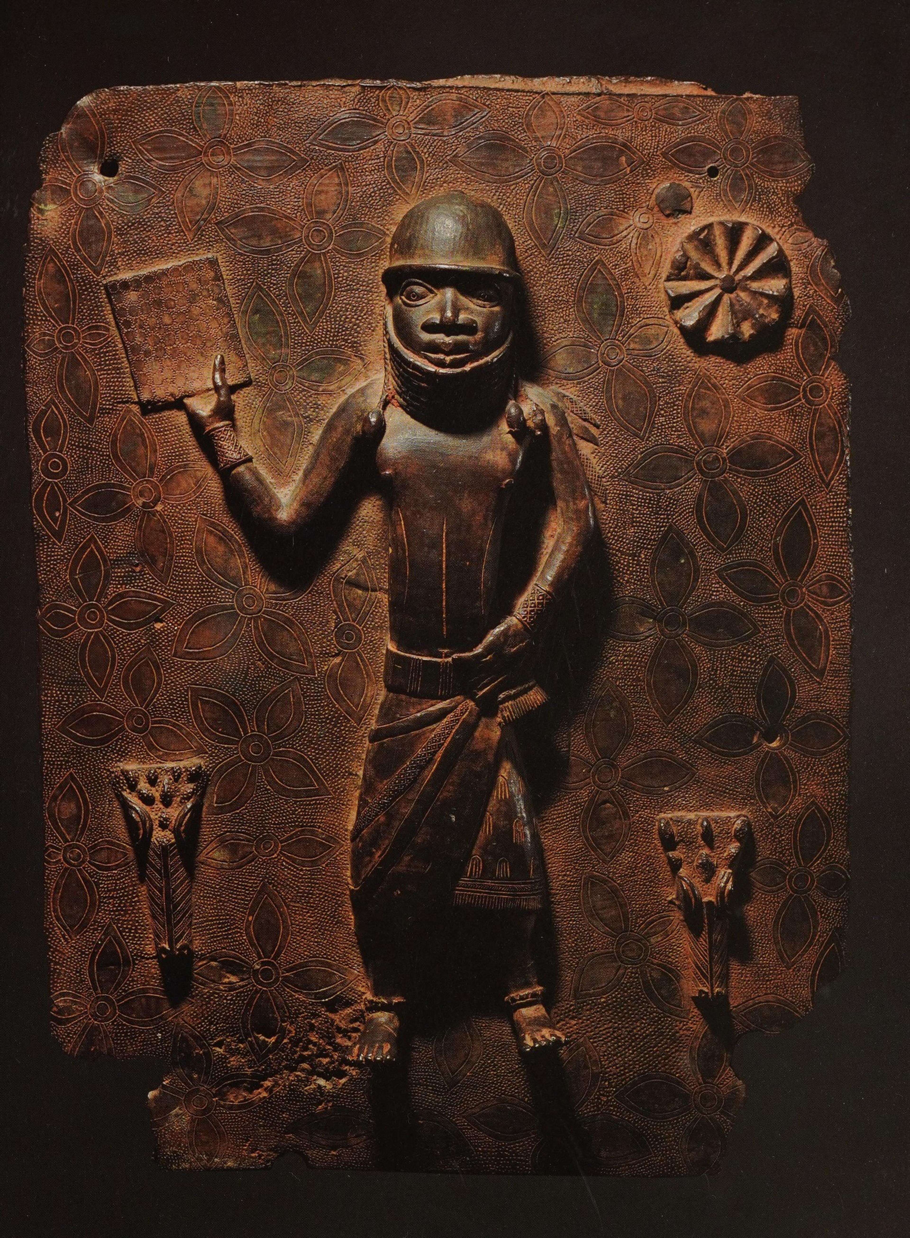 Bronze Benin sculpture with a figure holding a book. The background is adorned with floral motifs