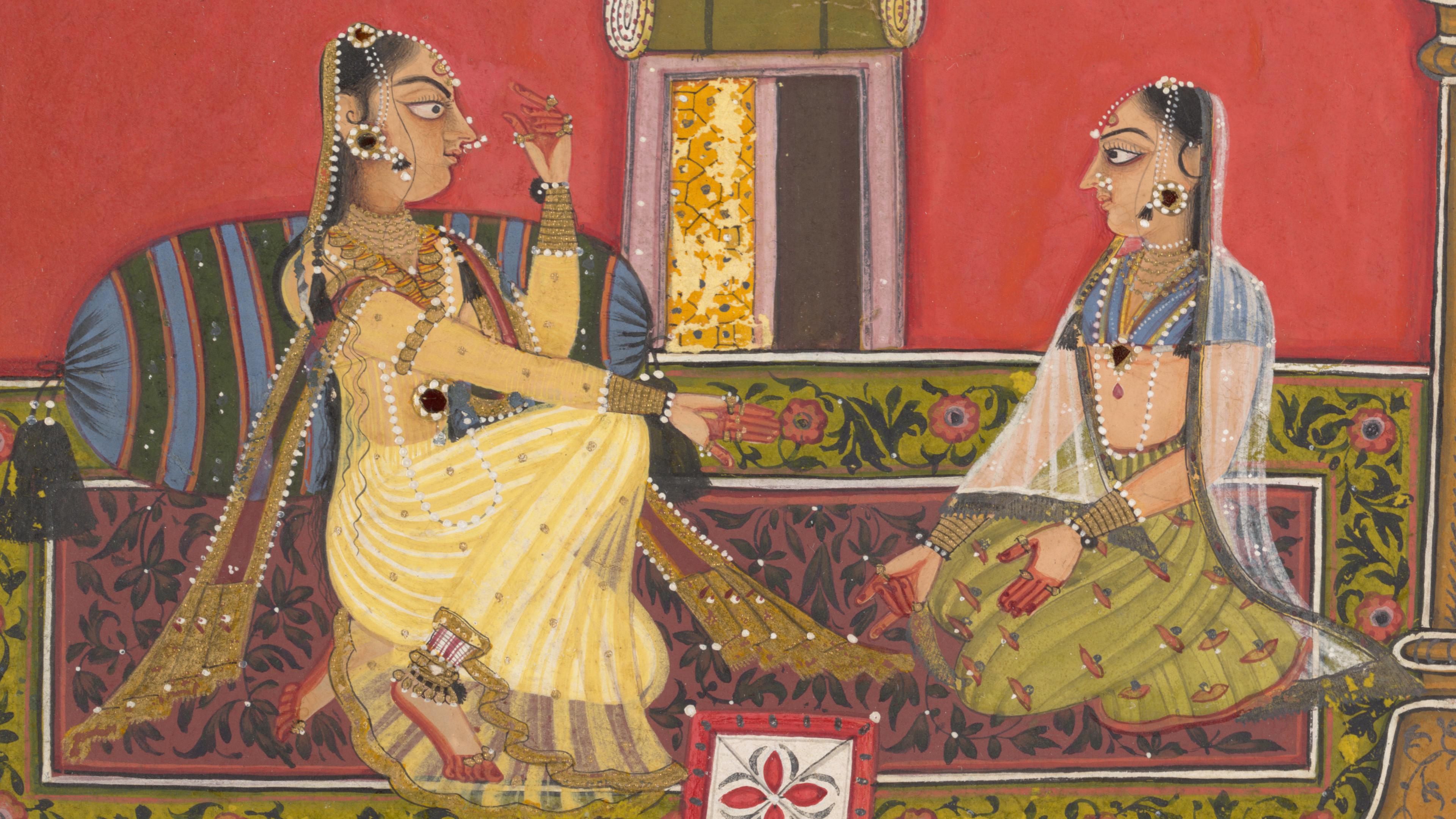 Two women in opulent dress and jewelry sit facing each other on a patterned carpet