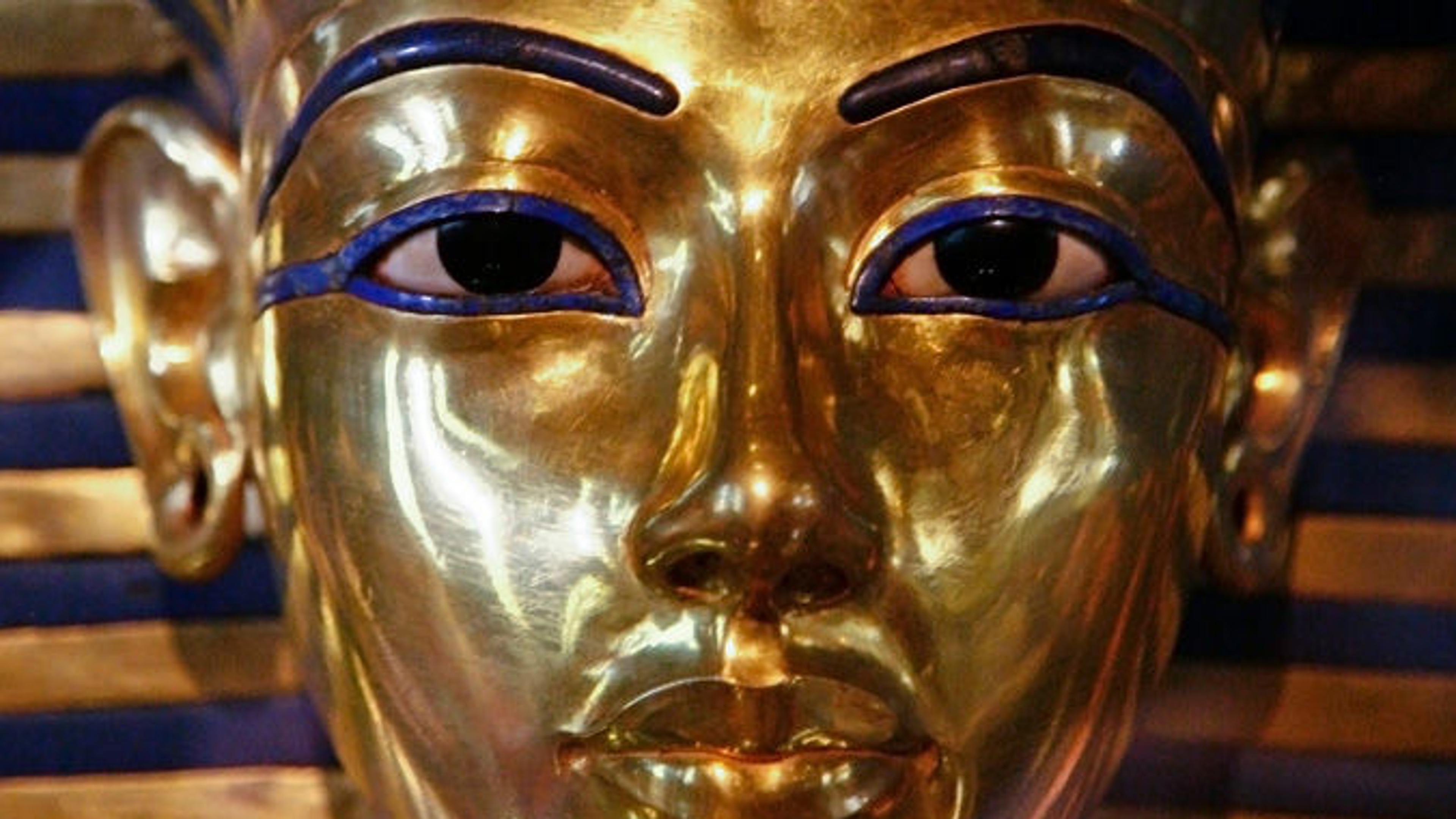 A gold mask with distinct facial features and a blue and gold stripe background.