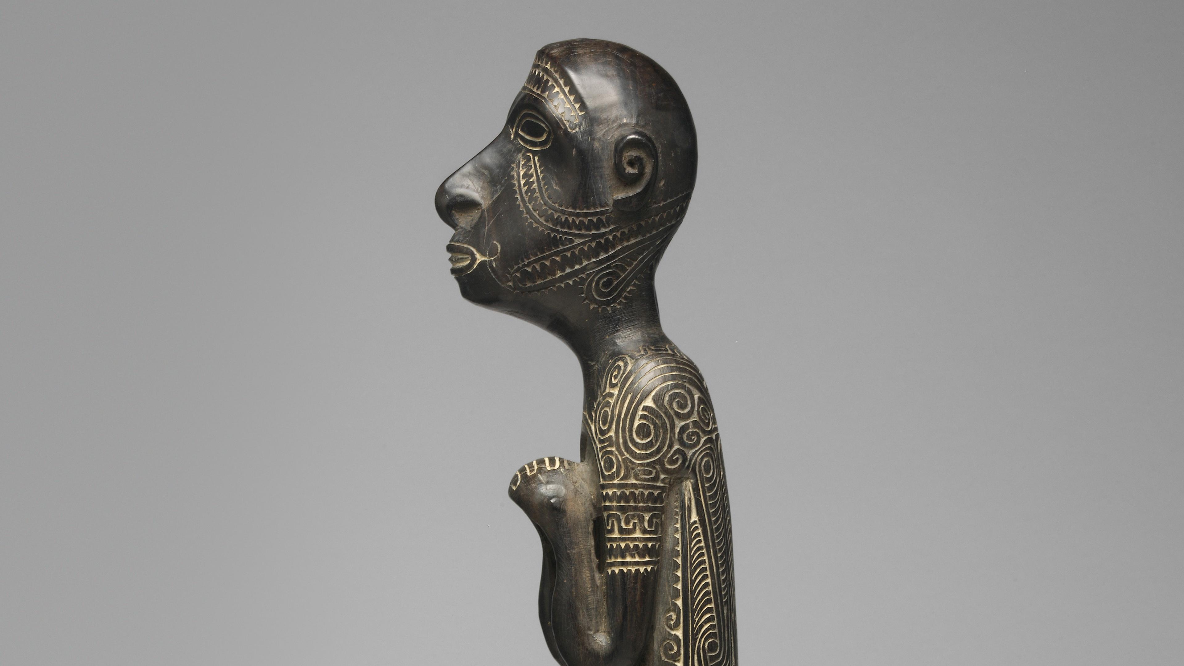 Detail of a carved human figure finial with hands clasped at its chest, with designs traced in white against the black surface.