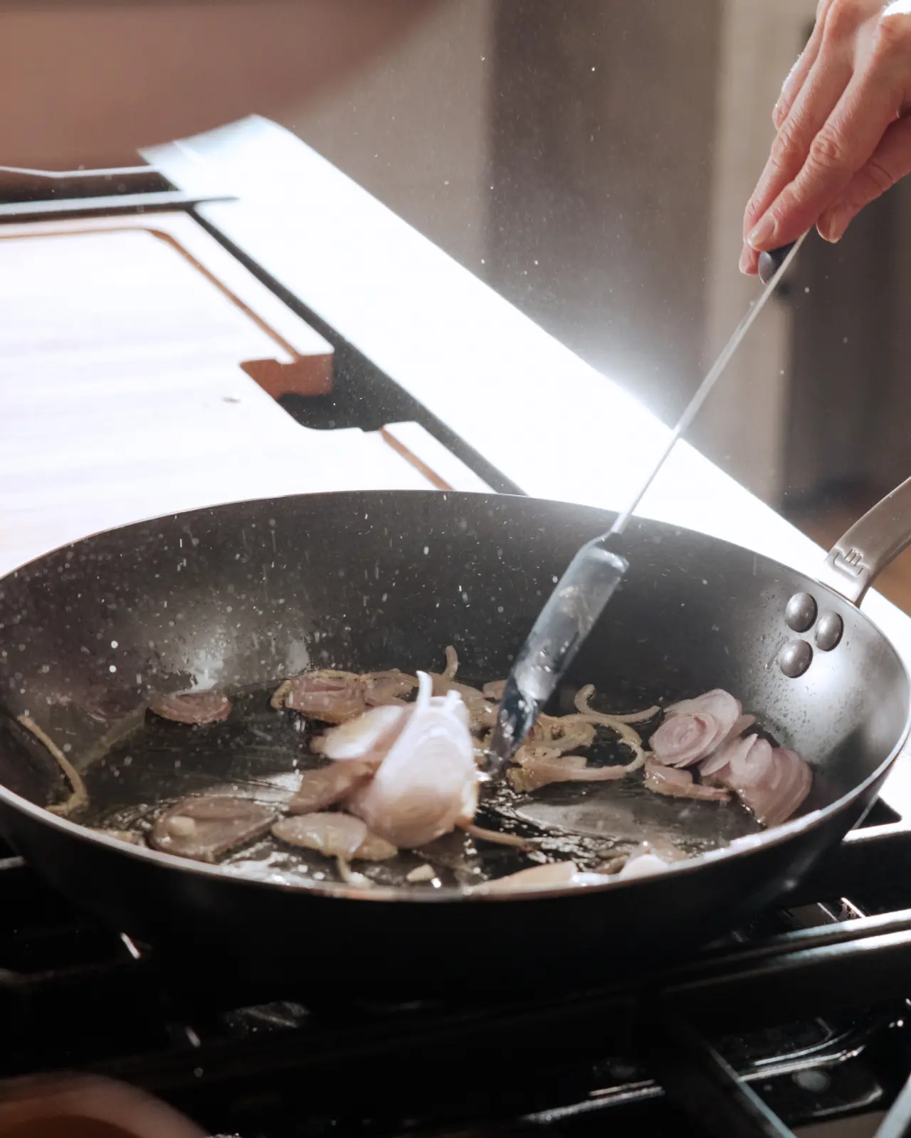 A hand stirs sliced onions sautéing in a pan, with steam rising from the cooking food.
