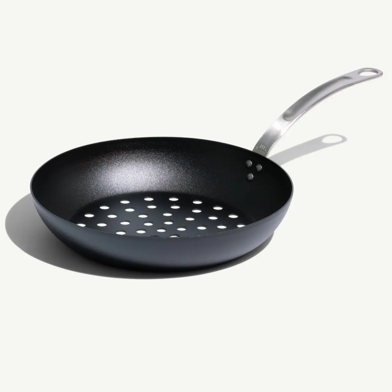 A black non-stick frying pan with a silver handle and multiple round holes in its base casts a shadow on a light surface.