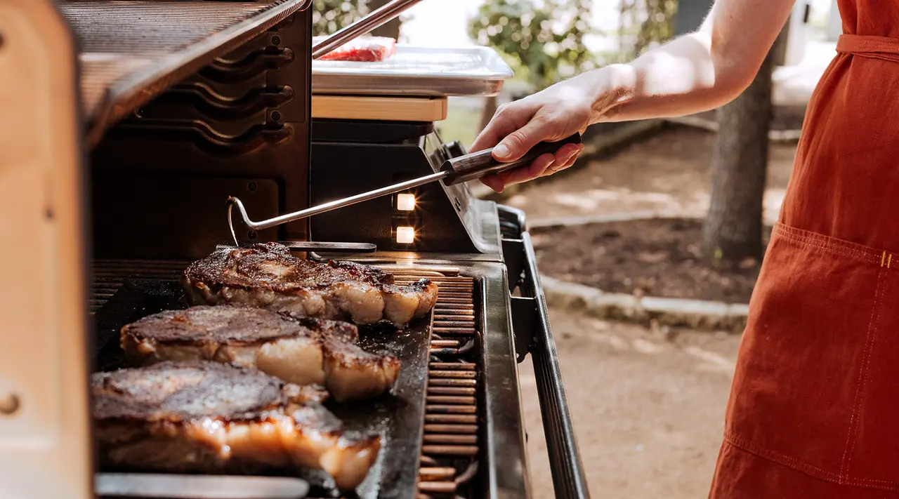 A person wearing a red apron grills steaks on a barbecue using long-handled tongs.