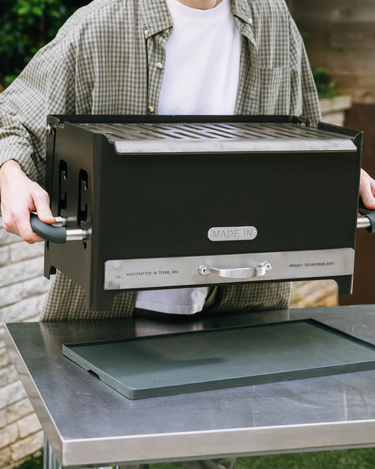 A person is carrying a black grill with handles, preparing to place it on a metal table outdoors.