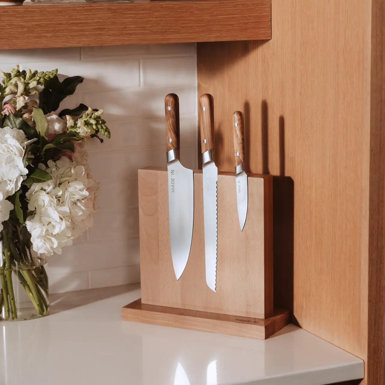 A wooden knife block holds three knives next to a bouquet of flowers on a countertop.
