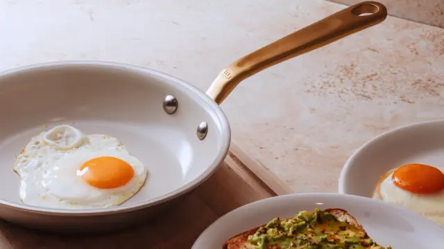 A white frying pan with a golden handle contains a sunny-side-up egg, accompanied by toasted bread topped with avocado and another egg.
