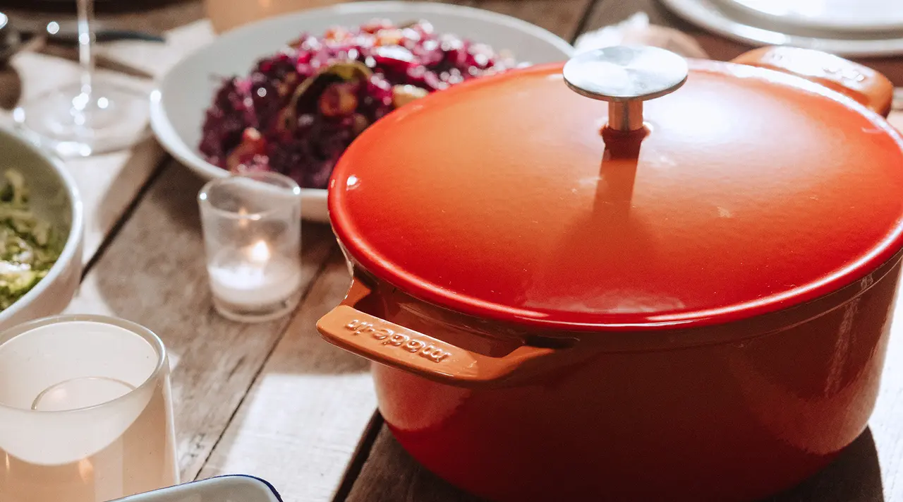 A vibrant orange cooking pot sits on a wooden table surrounded by dishes and candles.