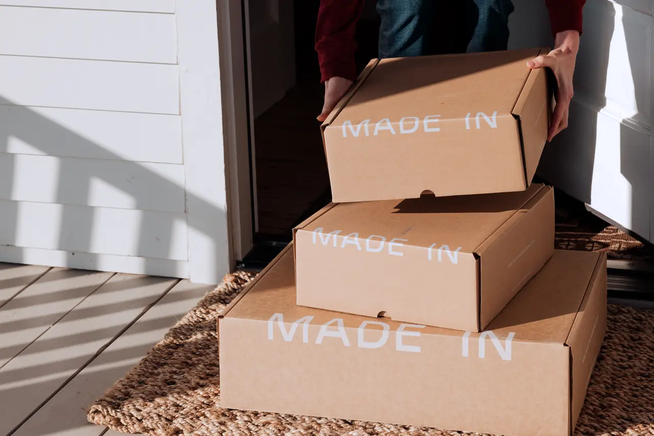 A person is holding a stack of cardboard boxes labeled "MADE IN," casting shadows in a sunlit room.
