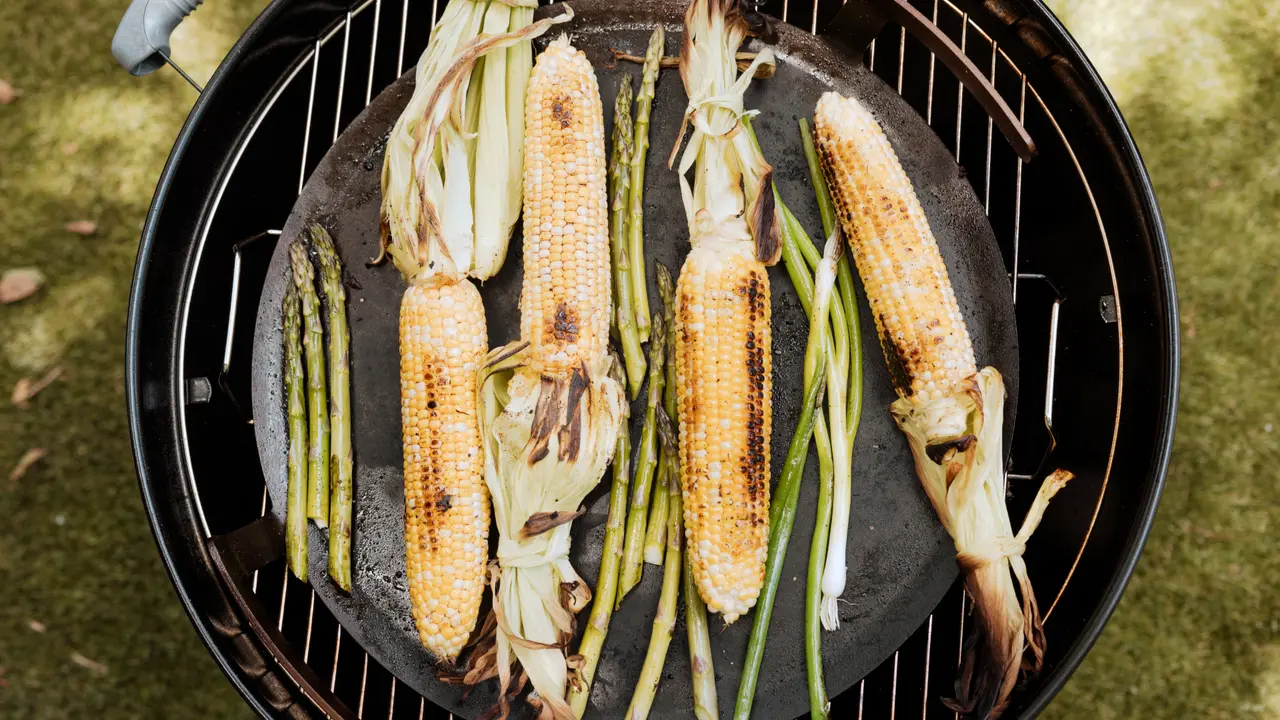 Grilled corn on the cob and green onions are cooking on a barbecue grill.