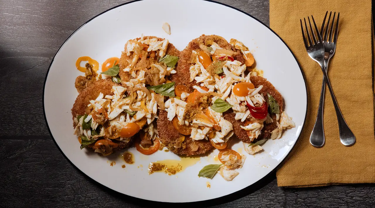 A plate of breaded chicken topped with cheese, vegetables, and sauce is accompanied by a fork and spoon on a dark table with a yellow napkin.
