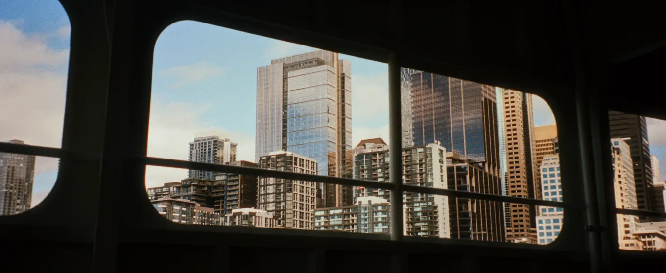 View of a cityscape through a ferry window, showcasing tall modern skyscrapers against a blue sky. The scene feels dynamic and urban.