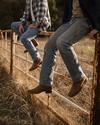 Two people wearing jeans and boots sit on a rusted metal fence in a grassy, rural outdoor setting. Their faces are not visible.