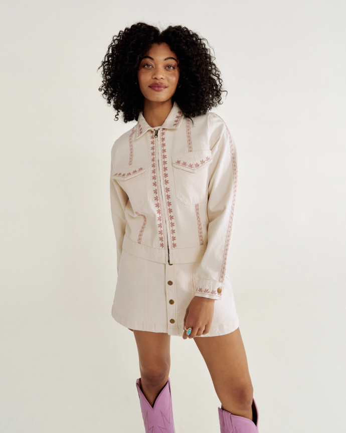 Person with curly hair wearing a light-colored jacket and skirt set with embroidery and pink cowboy boots, standing against a plain background.