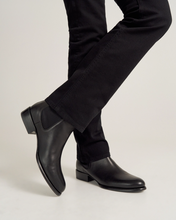 A person wearing black pants and black leather boots stands with one leg crossed over the other against a plain background.