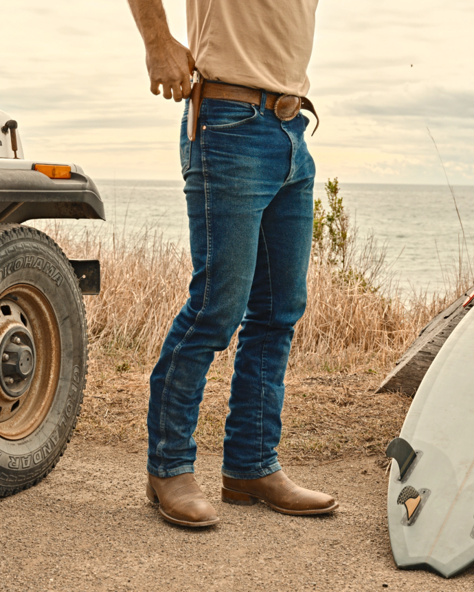 Person in jeans and boots stands by a coastal road beside a vehicle and surfboard, holding a phone.