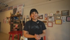 A person stands in a room with various framed pictures and artwork on the wall behind them, smiling at the camera and wearing an apron over a T-shirt. A red table and shelving are in the background.