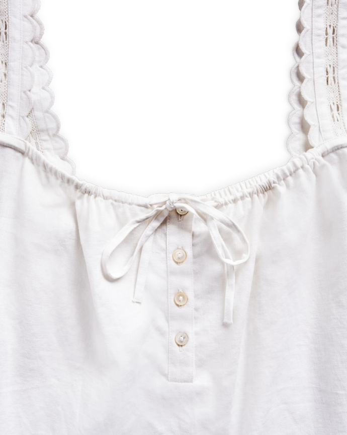 White sleeveless top with buttoned front, lace scalloped straps, and a bow tied at the neckline, against a black background.