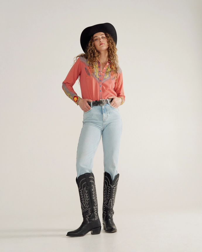 Person wearing a black cowboy hat, embroidered pink shirt, light blue jeans, and black cowboy boots stands against a plain background.
