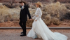 A bride and groom in western attire holding hands in a desert landscape, with the bride in a flowing gown and the groom in a black suit and cowboy hat.