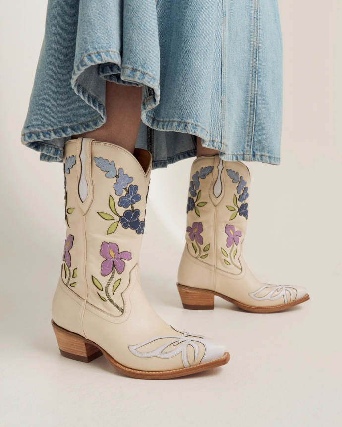 A person wearing a blue denim skirt and cream-colored cowboy boots with purple and blue floral embroidery.