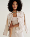 A woman with curly hair wears a light-colored jacket with embroidered patterns and a matching skirt. She has a turquoise necklace and stands against a plain background.
