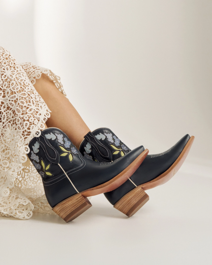 A person wearing a white lace dress and black cowboy boots with floral embroidery reclines with legs crossed on a light background.