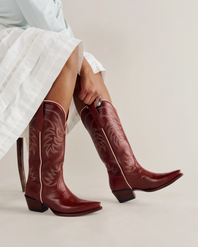 A person wearing a white skirt and red cowboy boots sits on a chair, with their hands resting on their legs against a plain background.