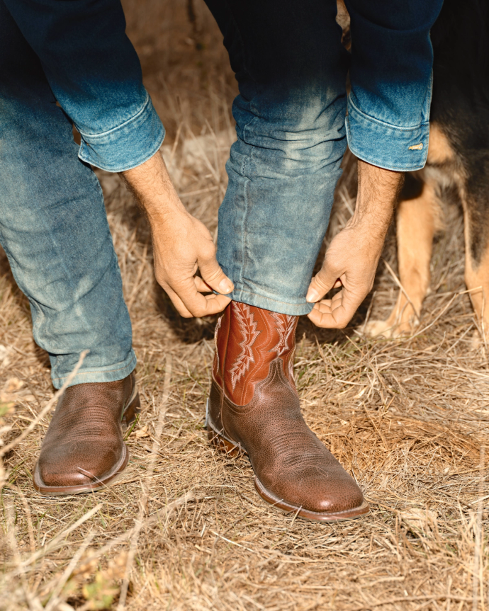A person wearing blue jeans adjusts their pant leg over brown cowboy boots while standing on dry grass.
