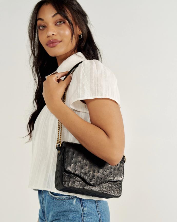 A woman in a white blouse and jeans holds a black textured handbag on her shoulder against a plain background.