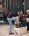 A man helps another man put on boots while others sit nearby, laughing and holding cans, in a casual setting.