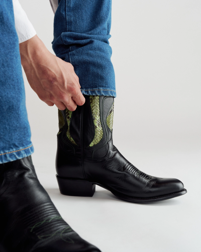 A person wearing blue jeans pulls on a black cowboy boot with green and black detail against a plain background.
