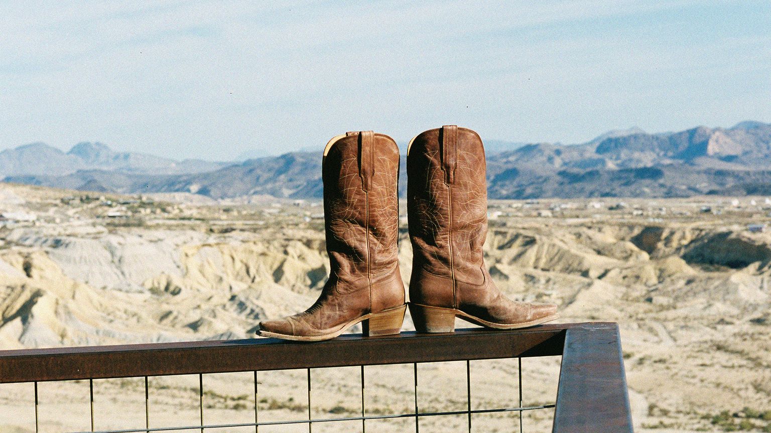 A pair of brown cowboy boots positioned on a fence with a vast desert landscape in the background.