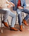 Two people sit on stools in a kitchen, holding drinks. They are dressed in denim jeans and brown boots, with one wearing a sweater and the other a denim jacket over a red shirt.