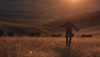 A person stands in a field at sunset, holding a hat and a walking stick, with cattle grazing in the background.