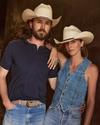 A man and woman, both wearing cowboy hats and denim clothing, stand closely together with relaxed expressions. The man has long hair and a beard, and the woman has her hand on his shoulder.