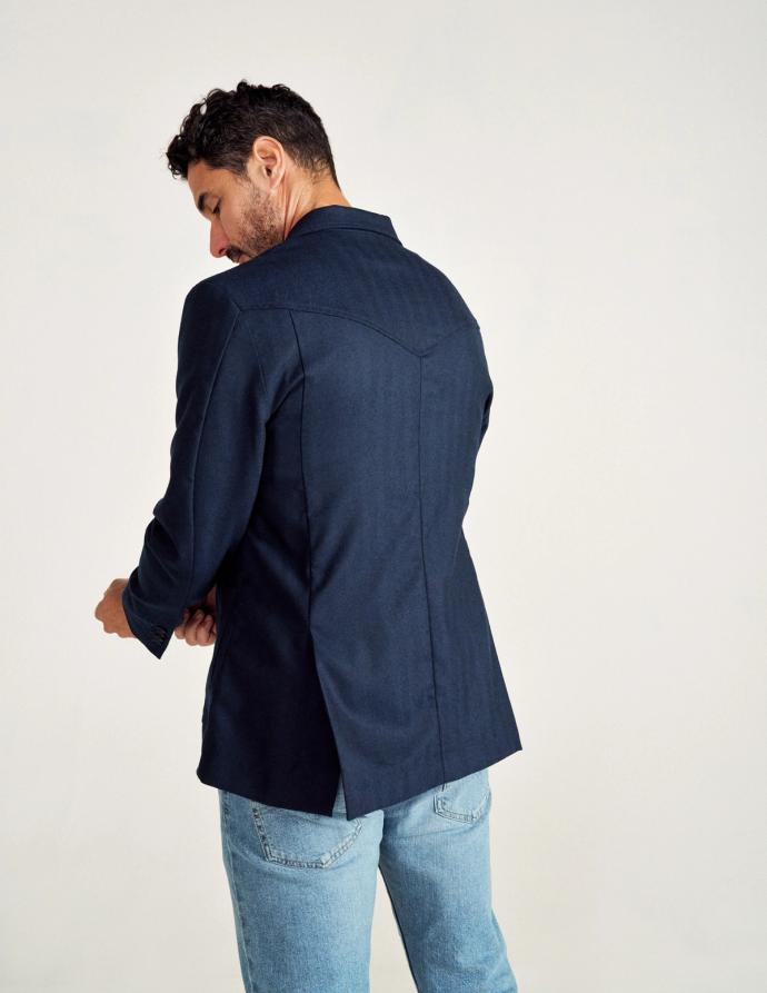 A man facing away, wearing a dark blazer and light blue jeans against a plain background.